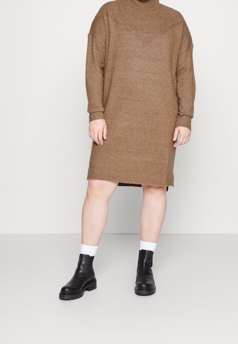 Robe pull marron en tricot avec col montant, coupe oversize et poignets côtelés. Portée avec des bottines noires épaisses et des chaussettes blanches.
