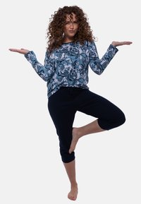 Pitkähihainen, kukkakuosiset paita sinisen sävyissä, yhdistetty tummiin capri-housuihin. Malli tasapainottelee yhdellä jalalla, paljain jaloin, tasaisella taustalla.