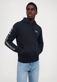 Felpa navy con tasca frontale e cappuccio. Presenta il logo EA7 sul petto e il testo del marchio lungo la manica. Tessuto morbido.