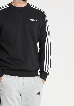 Mann trägt schwarzen Adidas-Sweatshirt mit weißen Streifen an den Ärmeln und hellgraue Adidas-Sweatpants mit Logo am Oberschenkel.