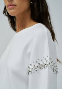 Sweatshirt blanc à col rond, orné de strass argentés décoratifs sur la manche, offrant un détail texturé et embellissant.