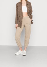 Beżowe spodnie dresowe z elastycznymi mankietami, z detalem logo. Połączone z brązową oversize'ową marynarką w kratę, białym topem crop i białymi sneakersami.