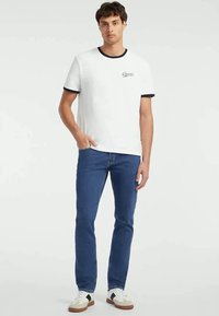 Jovem de pé com uma mão no bolso, vestindo uma t-shirt branca da Guess, jeans azuis e ténis brancos, num fundo simples.
