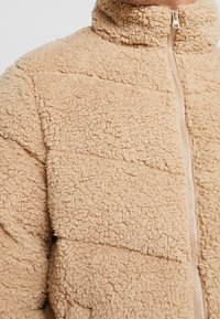 Beige fleece jack met een hoge kraag, featuring een ritssluiting en een gestructureerd, pluizig oppervlak met horizontale gestikte patronen.