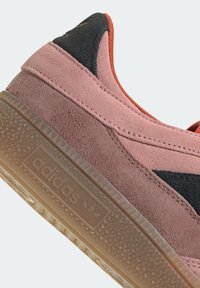 Adidas sneakers featuring pink suede upper, black accent, rounded toe, and textured gum rubber sole with prominent branding.
