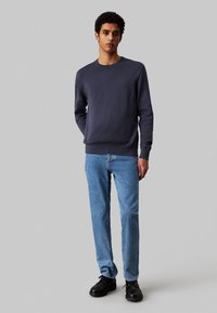 Felpa navy con colletto tondo, polsini e orlo a coste, abbinata a jeans blu chiaro e scarpe nere stringate. Design semplice, senza fantasie.