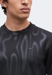 Άνδρας που φοράει μαύρη μπλούζα Nike με γυαλιστερά κυματιστά μοτίβα, φαίνεται από τους ώμους και πάνω, κοιτάζοντας ελαφρώς στο πλάι με φόντο ένα απλό φόντο.