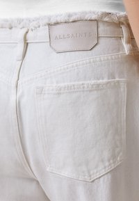 Jeans de mezclilla blanca con dobladillo deshilachado, que presentan una etiqueta de cuero marrón con la marca "ALLSAINTS" encima del bolsillo trasero.