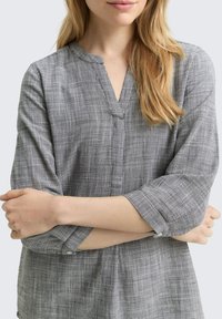 Mujer con cabello largo y rubio, vistiendo una blusa de manga larga a cuadros grises, con los brazos cruzados sobre el pecho, frente a un fondo claro.