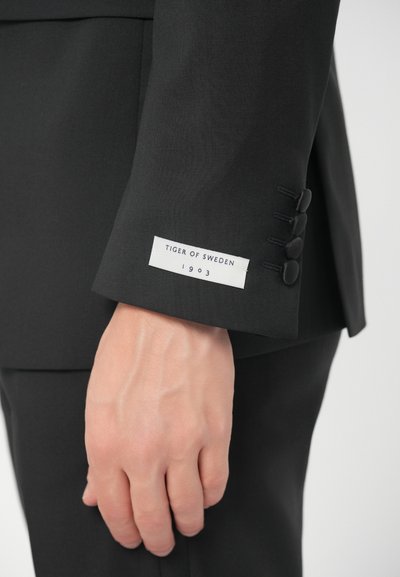 Ruka osoby vedle rukávu černého obleku s etiketou "Tiger of Sweden 1903" a čtyřmi černými knoflíky na manžetě.