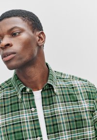 Green plaid shirt featuring vertical and horizontal lines in white, blue, and black, with a button-down collar and subtle logo detail.