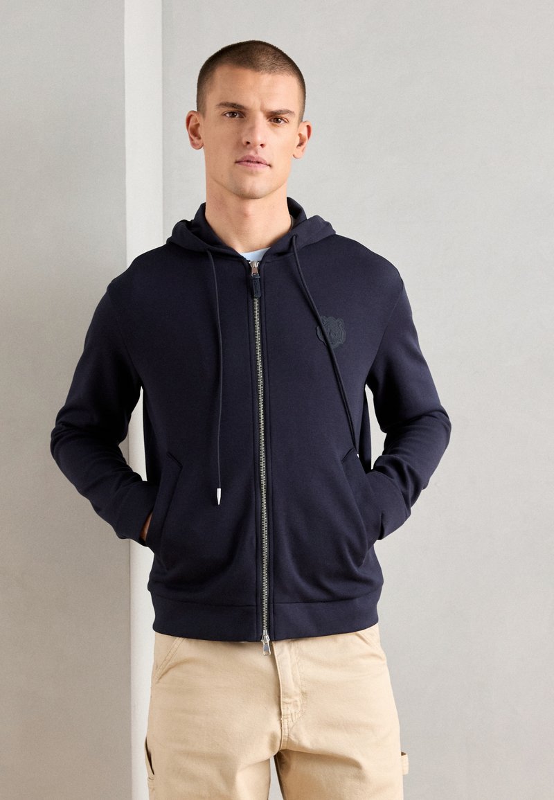 Marinefarbener Reißverschluss-Hoodie aus weichem Stoff mit Zugbändern und Fronttaschen. Prägiertes Logo auf der Brust. Schlicht, entspannter Schnitt.