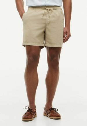 Mann trägt beige Shorts mit Kordelzug und braune Leder-Schnürschuhe, steht mit einer Hand in der Tasche.