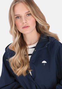 Tengerészkék vízálló dzseki, fehér esernyő logóval a mellkason, cipzáros kapucnival és légáteresztő hálós béléssel.