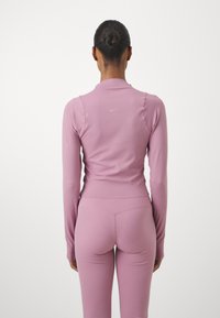 Kvinne med bun som har på seg en tettsittende mauve langermet topp og leggings, står med ryggen til mot en ensfarget hvit bakgrunn.