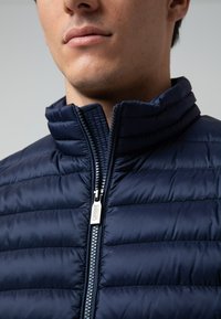 Giubbotto imbottito blu navy con zip frontale, dotato di colletto alto e motivo a cuciture orizzontali. Dettaglio della cerniera metallica visibile.