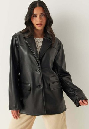 Kvinde med langt mørkt hår iført en sort læderblazer over en hvid skjorte med polkaprikker og beige bukser, stående foran en ensfarvet baggrund.