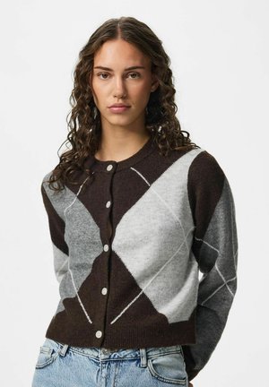 Jeune femme aux cheveux bouclés portant un cardigan boutonné marron et gris à motif losanges et un jean bleu clair sur fond uni.