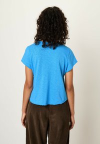 Modra kratka majica s kratkimi rokavi iz lahkega blaga, s krivim robom in surovim robom, v kombinaciji s smešnimi hlačami iz korduroja v rjavi barvi.