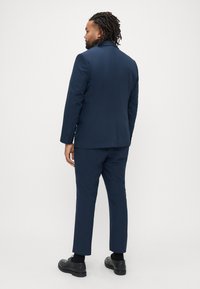 Marineblauw pak met een getailleerde pasvorm, voorzien van een enkellags colbert en smalle broek. Soepele stof en minimalistisch ontwerp. Zwarte schoenen.