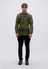 Εξωτερικό μπουφάν σε ελαιί πράσινο, ελαφρύ ύφασμα με τσέπη με φερμουάρ, συνδυασμένο με μαύρο παντελόνι και γκρι παπούτσια, που παρουσιάζει αθλητικό σχέδιο.