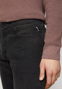 Črne jeanske hlače s oprijetim krojem, ki vključujejo diskretno šivanje, sprednje žepke in majhno črno oznako na zadnjem žepu.