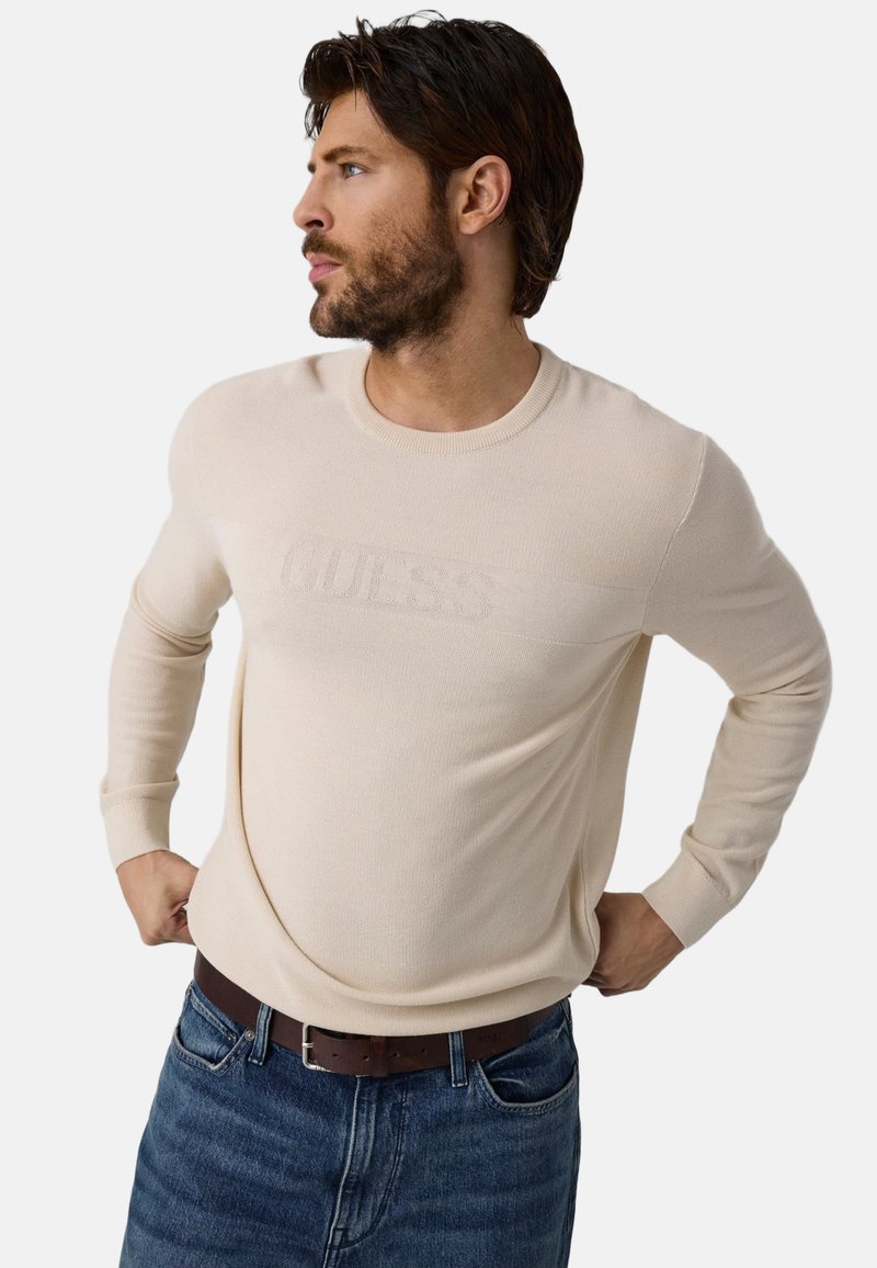 Άνδρας με γένια που φοράει μπεζ πουλόβερ Guess και μπλε τζιν, κοιτάζει αριστερά με τα χέρια στη μέση.