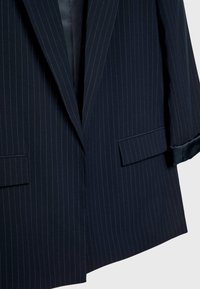 Laivastonsininen raidoitettu bleiseri, jossa on kauluskäänne, pitkät hihat ja läpätaskut. Ominaisuuksina sileä pinta ja satiinivuori.