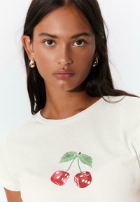 T-shirt bianco in cotone con una stampa colorata di due ciliegie e dadi con foglie verdi. Collo rotondo e maniche corte.