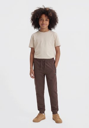 Enfant aux cheveux bouclés portant un t-shirt beige, un pantalon de jogging marron foncé et des baskets beige, debout avec une main dans la poche sur fond blanc.