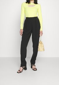 Keltainen pitkähihainen paita, jossa on aukko, mustat korkeavyötäröiset housut ja helmillä varustettu keltainen käsilaukku. Mustat paksut korot täydentävät asua.