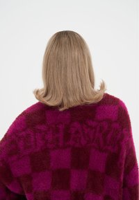 Měkký nadměrný svetr v kostkovaném vzoru v barvách bordó a magenta s jemnou texturou. Název značky je nenápadně začleněn do látky.