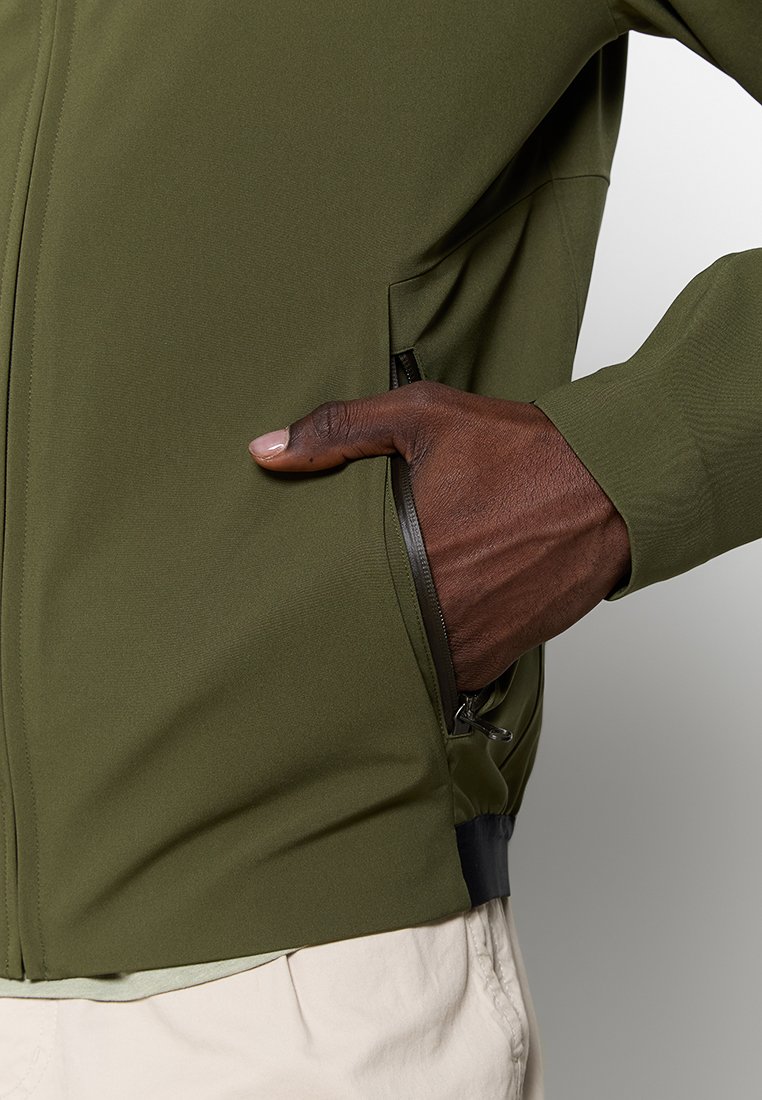 Mano nella tasca con zip di una giacca verde scuro, che mostra la texture della giacca e i pantaloni beige sottostanti.