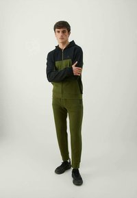 Haut à capuche zippé de couleur vert olive et noir avec un design bicolore et une silhouette ajustée. Porté avec des baskets noires.