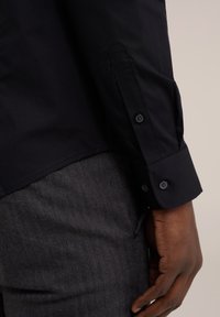 Pravá ruka v černé košili s dlouhým rukávem s knoflíkovým manžetou vedle šedých kalhot s herringbone vzorem, ukazující část ruky a kapsy.