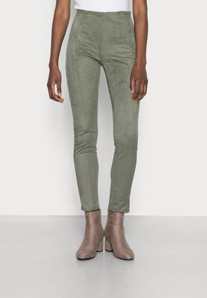 Slim-fit olivengrønne bukser båret med taupe ruskind ankelstøvler, vist på en person stående mod en ensfarvet lys baggrund.