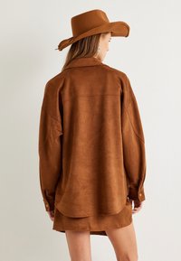 Chemise en suede marron à manches longues et col, avec un ourlet arrondi et des poignets à boutons. Assortie à un chapeau à large bord.