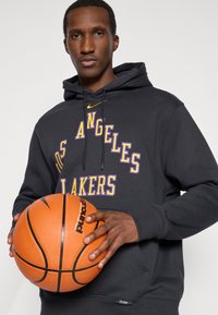 Juodas kapucinis su purpuriniu "Los Angeles Lakers" tekstu, reguliuojama virvute ir kengūros kišene, laikomas su oranžiniu krepšiniu rankose.