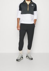 Chaqueta con cremallera en blanco y negro con detalles en naranja, combinada con pantalones jogger negros y zapatillas grises, con puños elásticos y tela texturizada.