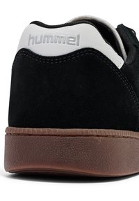 Schwarzer Wildlederschuh mit einem weißen Absatzakzent und "hummel"-Logo; strukturierte braune Gummisohle mit einem gemusterten Profil.