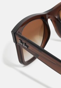 Ochelari de soare cu gradient maroniu, având un design masiv și unghiular. Prezintă un accent de ramă neagră și un logo Ray-Ban argintiu pe brățară.