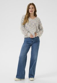 Jonge vrouw in een gespikkelde gebreide trui, met wijde blauwe jeans en witte sneakers, staande tegen een effen lichte achtergrond.