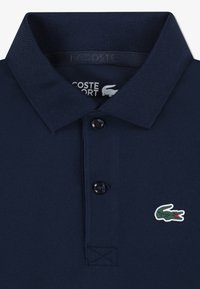 Tummansininen pikeepaita, joka on valmistettu strukturoidusta kankaasta. Siinä on kolmipainikkeinen aukko ja vihreä krokotiililogo vasemmassa rinnassa.