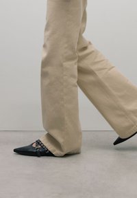 Zwarte puntige schoenen met enkelband en zilverkleurige hardware, gedragen met beige wijde broek, op een gladde grijze ondergrond.