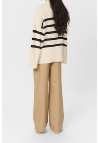 Femme aux longs cheveux noirs portant un pull crème avec des rayures noires et un pantalon large beige, debout devant un fond clair uni.