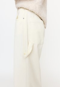 Zbliżenie na osobę w kremowych spodniach sztruksowych z boczną kieszenią i szlufką, w połączeniu z beżowym, teksturowanym swetrem.