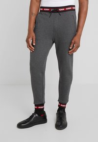 Graue Sweatpants mit slim Fit, elastischem Bund und einem roten und schwarzen Logodesign, kombiniert mit schwarzen Sneakers und Logosocken.