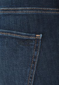 Närbild av baksidan av blå denimjeans med orange sömmar och en liten broderad fågel/logotyp.