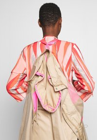 Henkilö, jolla on lyhyet kiharat hiukset, käyttää punaista, vaaleanpunaista ja valkoista kuviollista paitaa ja beigeä takkia, joka roikkuu hänen selästään.