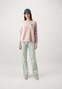 Felpa rosa chiaro con scollo rotondo e logo ricamato, abbinata a jeans azzurri e décolleté grigie a punta. Cappellino blu in testa.